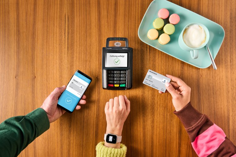 Ein Zahlgerät mit vielen Händen, die per Smartphone, Smartwatch oder Karte zahlen wollen