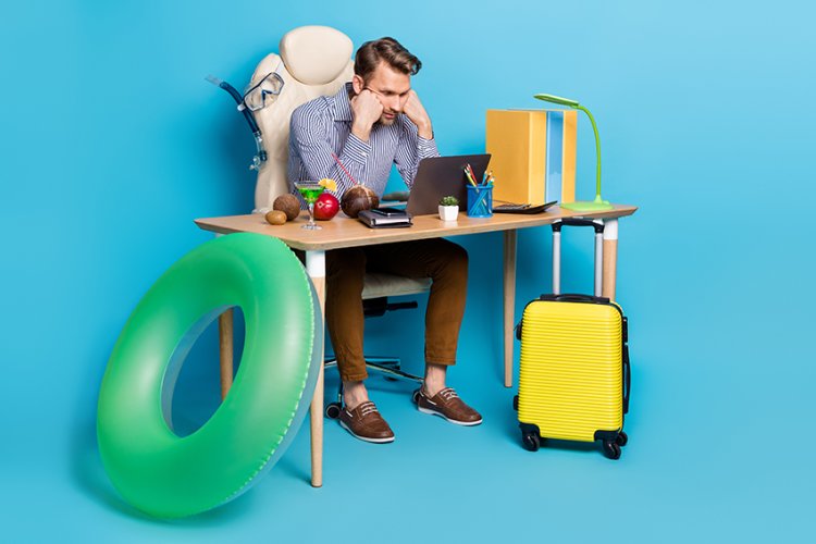 Ein Mann sitzt schlecht gelaunt am Schreibtisch, um ihn herum stehen Koffer